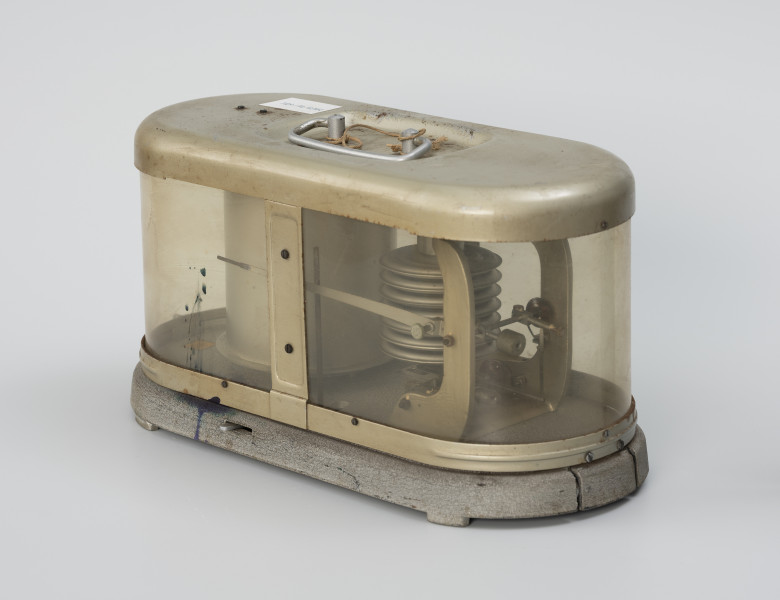 Barograf 207R - ujęcie z przodu ze skosu; Barograf typu 207R z puszką Vidiego i barogramem umieszczonymi na owalnej, metalowej podstawie, w obudowie z toczonej blachy, płaskowników i pleksi na ściankach bocznych. Na zewnętrznej części obudowy uchwyt do przenoszenia barografu. Puszka Vidiego składa się z 7 cylindrów umocowanych w ramie za pomocą elementu śrubowego z blokadą w postaci śrubki. Cylindryczny bęben, na którym umieszczany jest barogram, wyposażony jest od wewnątrz w zegar sprężynowy, którego napęd steruje obrotami bębna w równych odstępach czasu. W części wewnętrznej podstawy znajduje się tablica znamionowa.