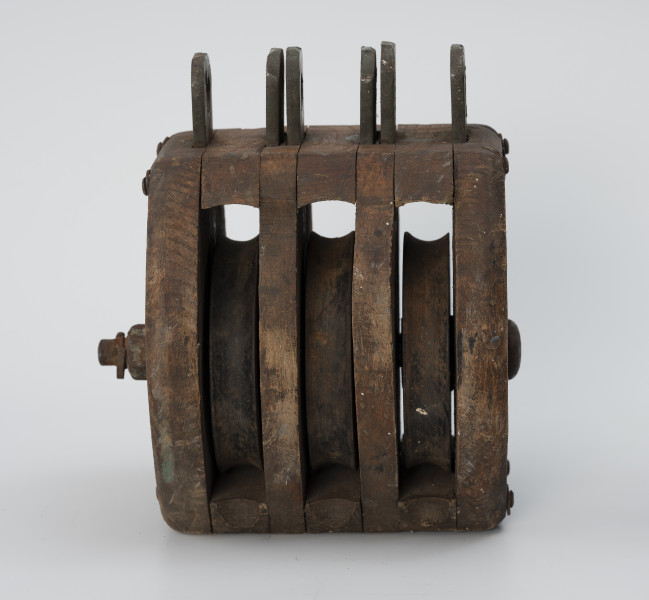 Blok okrętowy potrójny - ujęcie z tyłu; Blok drewniany potrójny, policzki owalne z zaokrąglonymi krawędziami o grubości ścianki 2,4 cm między policzkami a wstawami rozpórki o grubości 2,8 cm. Dwie wstawy drewniane o grubości 1,8 cm. Trzy krążki wykonane z metalu o średnicy 11 cm osadzone na osi wykonanej ze stali. Oś zabezpieczona sześciograniastą nakrętką (2,5 cm) z podkładką i przetyczką. Łeb osi okrągły o średnicy 3 cm, z podkładką. Zamocowany na stałe w poprzek policzków. Ucha stalowe składające się z osadzonych między policzkami i wstawami płaskowników długości 4 cm z otworami o średnicy 1,7cm. Zamocowano trzy zestawy uszu bez przetyczek. Policzki zespolone z rozpórkami i wstawami w całość czterema nitami stalowymi.