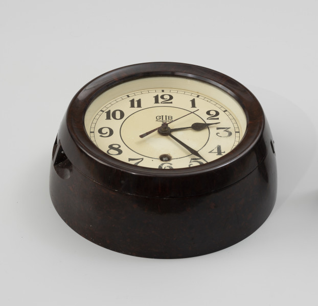 Zegar okrętowy - ujęcie z dołu; Mechaniczny zegar sprężynowy w okrągłej, bordowej obudowie z ebonitu z przeszkloną, okrągłą pokrywą, zamontowaną na zawiasie i zamykaną zamkiem na klucz kwadratowy. Ebonitową tarczę pomalowaną na jasnokremowy kolor przytwierdzona do płyty zegara. Powyżej cyfry z godziną 6. znajduje się okrągły otwór z kwadratowym zakończeniem wałka napędowego – zegar nakręcany jest za pomocą klucza kwadratowego. Tarcza wyskalowana na 12 godzin i 60 minut. Godziny oznaczone pełnymi cyframi arabskimi w kolorze czarnym umieszczone po wewnętrznej stronie tarczy. Pełne minuty zaznaczone prostokątem, wypełnionym czarnym kolorem. Wewnątrz tarczy barwą czarną obwiedzione koło o mniejszym promieniu, po środku umieszczono walcowy czop, na którym znajdują się wskazówki, z blachy barwionej na czarno – wskazówki godzinowa i minutowa i na bordowo – sekundowa. Najkrótsza wskazówka godzinowa sięga do podziałki godzinowej, a minutowa do podziałki minutowej, Na wierzchu czopu umieszczona jest wskazówka sekundowa – cienka i prosta strzałka z przeciw ciężarem. W górnej części wewnętrznej strony tarczy umieszczony napis GUB Glashutte/SA.