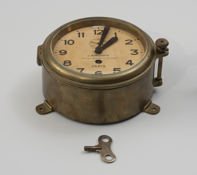 Zegar okrętowy - ujęcie z dołu; Zegar okrętowy w metalowej, okrągłej obudowie z sygnaturą J. Auricoste a Paris od wewnętrznej strony. Tarcza w kolorze kremowym o średnicy 14,5 cm. Podziałka godzinowo-minutowa, na której cyframi arabskimi od 1 do 12 oznaczono jedynie godziny. Wskazówki godzinowa i minutowa umieszczone centralnie, kształt prosty, bez ozdób. Tarcza sekundowa ponad osią, pomiędzy centrum tarczy a liczbą 12, z podziałką wyskalowaną od 0 do 60 sekund, na której podpisano wartość co dziesiątej sekundy. Wskazówka sekundowa czarna prosta. Pod osią trzpień do nakręcania o kwadratowym przekroju. Pomiędzy trzpieniem a osią godzinową znajduje się sygnatura J. Auricoste - horloger de la Marine de l'Elat - Paris. Średnica obudowy zegara wynosi 18 cm, wysokość 8,5 cm. Pokrywa szklana w obudowie pierścieniowej, profilowanej, z uszczelką gumową o zawiasowym zaczepie, zamykana na trzpień i ozdobną śrubę. Na obudowie od spodu uchwyty do mocowania zegara do podłoża. Klucz o motylkowatym uchwycie i przekroju wewnętrznym kwadratowym 3,5 x 3,5 mm.