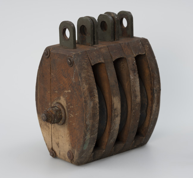 Blok okrętowy potrójny - ujęcie ze skosu z lewej strony; Blok drewniany potrójny, policzki owalne z zaokrąglonymi krawędziami o grubości ścianki 2,4 cm między policzkami a wstawami rozpórki o grubości 2,8 cm. Dwie wstawy drewniane o grubości 1,8 cm. Oś zabezpieczona sześciograniastą nakrętką (2,5 cm) z podkładką i przetyczką. Łeb osi okrągły o średnicy 3 cm, z podkładką. Zamocowany na stałe w poprzek policzków. Ucha stalowe składające się z osadzonych między policzkami i wstawami płaskowników długości 4 cm z otworami o średnicy 1,7cm. Zamocowano trzy zestawy uszu bez przetyczek. Policzki zespolone z rozpórkami i wstawami w całość czterema nitami stalowymi.