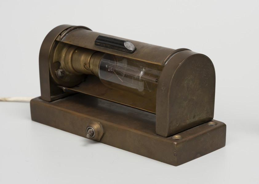 Elektryczna lampa kabinowa - ujęcie ze skosu; Lampa elektryczna z korpusem w całości zrobionym z mosiężnej blachy o prostokątnej podstawie (193 x 70 mm, wys 18mm) i z włącznikiem umieszczonym od czoła. Od góry, w podstawie cztery otwory do mocowania lampy, a na podstawie przymocowane dwie ścianki (12 x 60, wys. 70) zaokrąglone od góry. Pomiędzy ściankami znajduje się ruchoma przesłona umożliwiająca zmianę kąta padania światła. Na przesłonie umocowano czarny uchwyt z tworzywa sztucznego. Żarówkę (moc 66 W, światłość 26 cd, typ trzonka B22) wyprodukowano na terenie byłej NRF w firmie 