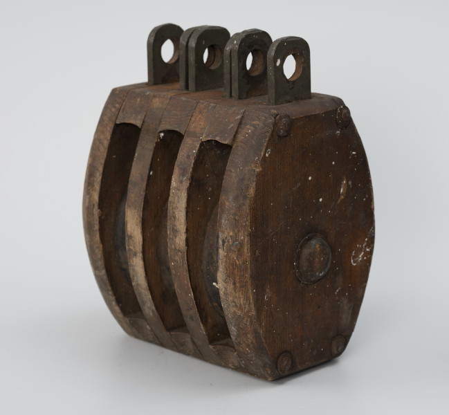 Blok okrętowy potrójny - ujęcie ze skosu z prawej strony; Blok drewniany potrójny, policzki owalne z zaokrąglonymi krawędziami o grubości ścianki 2,4 cm. Dwie wstawy drewniane o grubości 1,8 cm. Ucha stalowe składające się z osadzonych między policzkami i wstawami płaskowników długości 4 cm z otworami o średnicy 1,7cm. Zamocowano trzy zestawy uszu bez przetyczek. Policzki zespolone z rozpórkami i wstawami w całość czterema nitami stalowymi.
