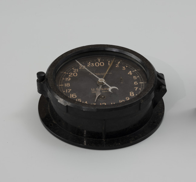 Zegar okrętowy - ujęcie z dołu; Czarna, ebonitowa obudowa w kształcie walca. Spodnia część wydłużona z trzema otworami do montowania na ścianie. Pokrywa z plastiku wykonana jako przeszklony pierścień montowany na zawiasie z prawej strony, z lewej strony gwintowana śruba zamykająca pierścień, szkło wykonane z przeźroczystego plastiku. Pod pokrywą tarcza z czarnego plastiku przykręcona do obudowy czterema śrubami. Na tarczy 24-godzinna biała skala zegarowa z białymi cyframi arabskimi, po wewnętrznej stronie godziny, od 00 do 23, po zewnętrznej minuty opisane liczbowo wartościami co 5 od 5 do 60. Na dole tarczy, powyżej liczby 12 znajduje się okrągły otwór z kwadratowym zakończeniem wałka napędowego (zegar nakręcany za pomocą klucza kwadratowego). W centrum tarczy czop z osadzonymi metalowymi wskazówkami, godzinowa w kształcie wskaźnika, minutowa strzałki, sekundowa prosta z zakończeniem w kształcie półksiężyca. Nad czopem wskazówkowym napis Chelsea Clock Co. Boston, a poniżej napis U.S. NAVY ser. no. 62415E. Po prawej str. na wysokości godzin 5. i 6. otwór prostokątny z okrągłym suwadłem do regulacji szybkości chodu zegara. Nad otworem litera F, pod litera S.