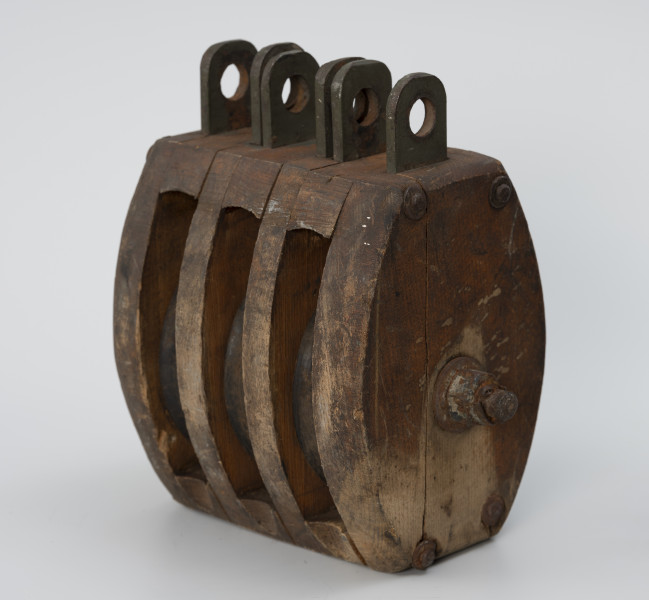 Blok okrętowy potrójny - ujęcie ze skosu z prawej strony; Blok drewniany potrójny, policzki owalne z zaokrąglonymi krawędziami o grubości ścianki 2,4 cm. Dwie wstawy drewniane o grubości 1,8 cm. Oś zabezpieczona sześciograniastą nakrętką (2,5 cm) z podkładką i przetyczką. Łeb osi okrągły o średnicy 3 cm, z podkładką. Zamocowany na stałe w poprzek policzków. Ucha stalowe składające się z osadzonych między policzkami i wstawami płaskowników długości 4 cm z otworami o średnicy 1,7cm. Zamocowano trzy zestawy uszu bez przetyczek. Policzki zespolone z rozpórkami i wstawami w całość czterema nitami stalowymi.