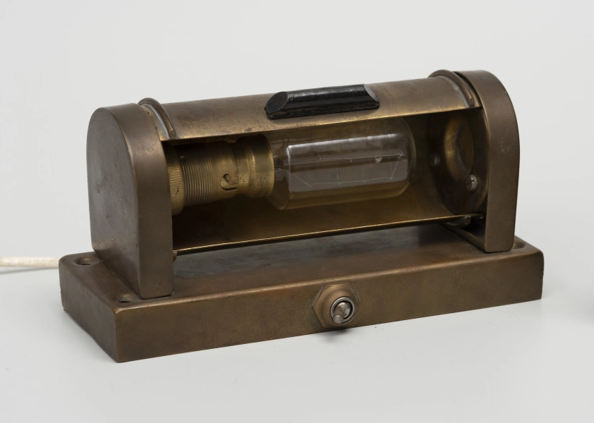 Elektryczna lampa kabinowa - ujęcie ze skosu z drugiej strony; Lampa elektryczna z korpusem w całości zrobionym z mosiężnej blachy o prostokątnej podstawie (193 x 70 mm, wys 18mm) i z włącznikiem umieszczonym od czoła. Od góry, w podstawie cztery otwory do mocowania lampy, a na podstawie przymocowane dwie ścianki (12 x 60, wys. 70) zaokrąglone od góry. Pomiędzy ściankami znajduje się ruchoma przesłona umożliwiająca zmianę kąta padania światła. Na przesłonie umocowano czarny uchwyt z tworzywa sztucznego. Żarówkę (moc 66 W, światłość 26 cd, typ trzonka B22) wyprodukowano na terenie byłej NRF w firmie 