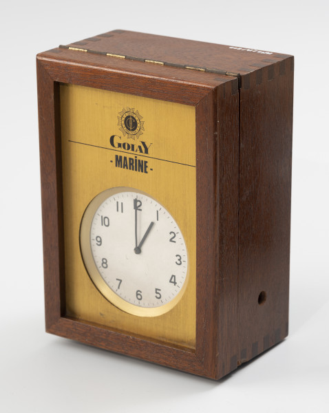 Elektroniczny zegar okrętowy - ujęcie ze skosu; Elektroniczny zegar okrętowy marki GolaY – Marine w obudowie z lakierowanego drewna. Pokrywa obudowy na zawiasie otwierana do góry. W mosiężnej pokrywie zegara znajduje się okrągła tarcza w kolorze białym, na niej w kolorze czarnym naniesiono skalę godzinową, oznaczoną pogrubionymi kreskami podziałki i cyframi arabskimi od 1 do 12 oraz skalę minutowo-sekundową, oznaczoną cieńszymi kreskami. Wskazówka minutowa i godzinowa wykonane z metalu, proste, w kolorze czarnym, brak wskazówki sekundowej. Tarcza zabezpieczona szkłem. Powyżej tarczy, na mosiężnym froncie obudowy widoczna sygnatura w kształcie rozety z okrągłym polem w centrum z napisem SWISS PRECISION i 10 gwiazdkami w otoku, centrum pola czarne z negatywowym symbolem długości geograficznej. Pod rozetą czarny napis w dwóch wierszach: Golay - MARINE -.