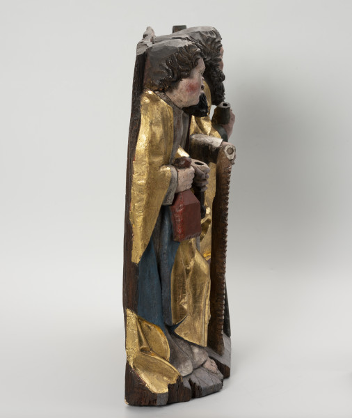 grupa rzeźbiarska - ujęcie z lewej; Płaskorzeźbiona płyta o kształcie stojącego prostokąta. Polichromowana i złocona. Przedstawia grupę trzech apostołów stojących na szerokiej podstawie. Zewnętrze postaci wysunięte są do przodu. Pomiędzy nimi, widoczny częściowo, stoi trzeci z apostołów. Ubrani są w tuniki i obszerne, plastycznie modelowane szaty. Św. Bartłomiej ukazany jako młody mężczyzna o gładkim licu trzyma w dłoniach atrybuty. Pozostali apostołowie noszą długie kędzierzawe brody. Przed sobą trzymają atrybuty: św. Szymon - piłę. Ubytki drewna widoczne są w podstawie. Tył płyty opracowany płasko.