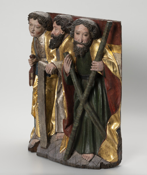 grupa rzeźbiarska - ujęcie ze skosu z prawej; Płaskorzeźbiona płyta o kształcie stojącego prostokąta. Polichromowana i złocona. Przedstawia grupę trzech apostołów stojących na szerokiej podstawie na tle kotary z frędzlami. Zewnętrze postaci wysunięte są do przodu. Pomiędzy nimi, widoczny częściowo, stoi trzeci z apostołów. Ubrani są w tuniki i obszerne, plastycznie modelowane szaty. Św. Bartłomiej ukazany jako młody mężczyzna o gładkim licu trzyma w dłoniach atrybuty. Ostrze noża nie zachowało się. Pozostali apostołowie noszą długie kędzierzawe brody. Przed sobą trzymają atrybuty: św. Szymon - piłę, a św. Andrzej - krzyż o kształcie litery 