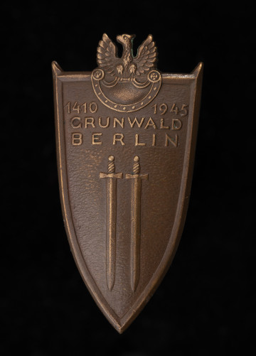 MNS/H/732/3 Odznaka Grunwaldzka - ujęcie z przodu; Brązowa odznaka w kształcie tarczy normańskiej, ale z płaską górną krawędzią. Pole odznaki gładkie, lekko wypukłe, krawędzie szerokie, podniesione, w narożnikach u góry wychodzące w formie szpiców poza tarczę. W polu wypukłe dwa miecze zwrócone głowniami w dół, nad nimi napis w 3 wierszach: 1410 1945 / GRUNWALD / BERLIN. Zwieńczeniem odznaki jest orzeł wojskowy siedzący na górnej krawędzi tarczy Amazonek, umieszczonej w górnej części odznaki. Korpus orła wystaje nad tarczę odznaki - głowa mała, szyja i korpus pokryte