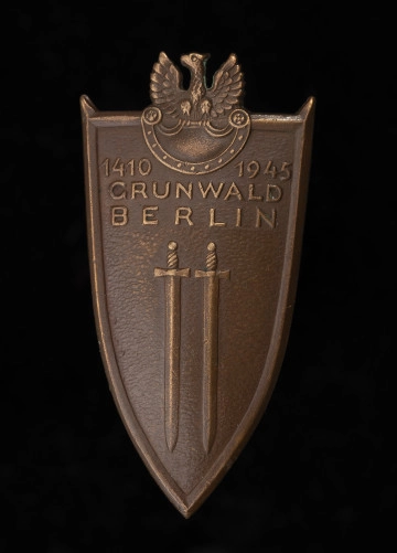 MNS/H/732/3 Odznaka Grunwaldzka - ujęcie z przodu; Brązowa odznaka w kształcie tarczy normańskiej, ale z płaską górną krawędzią. Pole odznaki gładkie, lekko wypukłe, krawędzie szerokie, podniesione, w narożnikach u góry wychodzące w formie szpiców poza tarczę. W polu wypukłe dwa miecze zwrócone głowniami w dół, nad nimi napis w 3 wierszach: 1410 1945 / GRUNWALD / BERLIN. Zwieńczeniem odznaki jest orzeł wojskowy siedzący na górnej krawędzi tarczy Amazonek, umieszczonej w górnej części odznaki. Korpus orła wystaje nad tarczę odznaki - głowa mała, szyja i korpus pokryte