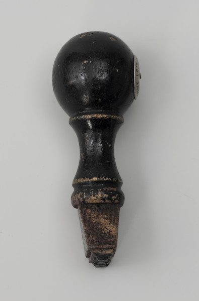 pieczątka firmowa - Ujęcie z boku; Pieczątka z napisem do odbijania, wykonanym minuskułą typu gotyckiego: Napis wycięty w wąskim prostokątnym kawałku tworzywa sztucznego (prawdopodobnie linoleum) i przyklejony do drewnianego uchwytu zakończonego okrągłą główką, na której przymocowano za pomocą nitu metalowy znak firmowy.  Od główki odchodzi wąska szyjka, do której przytwierdzony jest element o przekroju w kształcie trapezu. Drewniana część tłocząca zabarwiona czarnym lakierem.