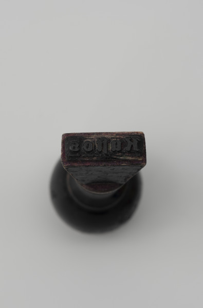 pieczątka firmowa - Ujęcie z dołu; Pieczątka z napisem do odbijania. Kustos - napis do góry nogami wycięty w wąskim prostokątnym kawałku tworzywa sztucznego (prawdopodobnie linoleum) i przyklejony do drewnianego uchwytu.