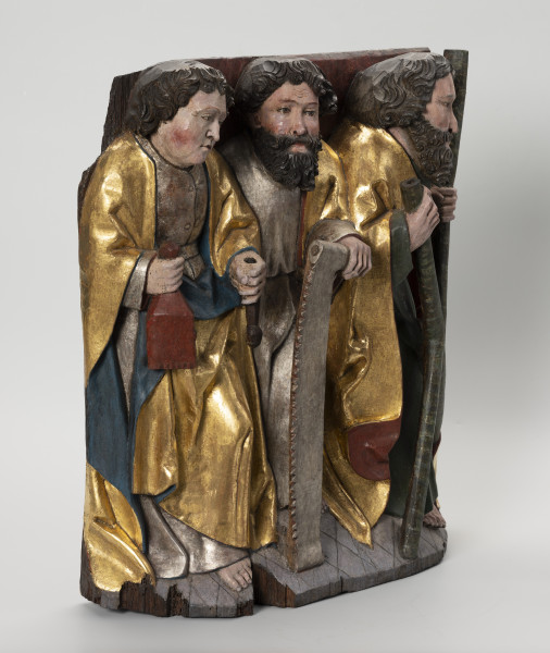 grupa rzeźbiarska - ujęcie ze skosu z lewej; Płaskorzeźbiona płyta o kształcie stojącego prostokąta. Polichromowana i złocona. Przedstawia grupę trzech apostołów stojących na szerokiej podstawie na tle kotary z frędzlami. Zewnętrze postaci wysunięte są do przodu. Pomiędzy nimi, widoczny częściowo, stoi trzeci z apostołów. Ubrani są w tuniki i obszerne, plastycznie modelowane szaty. Św. Bartłomiej ukazany jako młody mężczyzna o gładkim licu trzyma w dłoniach atrybuty. Ostrze noża nie zachowało się. Pozostali apostołowie noszą długie kędzierzawe brody. Przed sobą trzymają atrybuty: św. Szymon - piłę, a św. Andrzej - krzyż o kształcie litery 