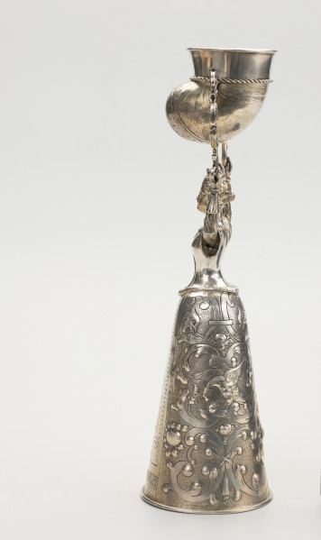 puchar weselny - ujęcie z lewej; Puchar w kształcie młodej kobiety w renesansowej sukni z mocno zasznurowanym staniczkiem i spódnicą o stożkowym kształcie, zdobioną ornamentem schweifwerkowym przechodzącymi w liście akantu, motywami ptaków i kiści owoców. Włosy dziewczyny są spięte z tyłu w koczek, na głowie nosi niewielką koronę z czterema kwiatonami. W rękach wzniesionych nad głową trzyma parę ażurowych wolutowych wsporników, pomiędzy którymi na trzpieniach zamontowany został pucharek w kształcie muszli.