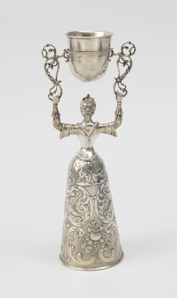 puchar weselny - ujęcie z tyłu; Puchar w kształcie młodej kobiety w renesansowej sukni z mocno zasznurowanym staniczkiem i spódnicą o stożkowym kształcie, zdobioną ornamentem schweifwerkowym przechodzącymi w liście akantu, motywami ptaków i kiści owoców. Włosy dziewczyny są spięte z tyłu w koczek, na głowie nosi niewielką koronę z czterema kwiatonami. W rękach wzniesionych nad głową trzyma parę ażurowych wolutowych wsporników, pomiędzy którymi na trzpieniach zamontowany został pucharek w kształcie muszli.