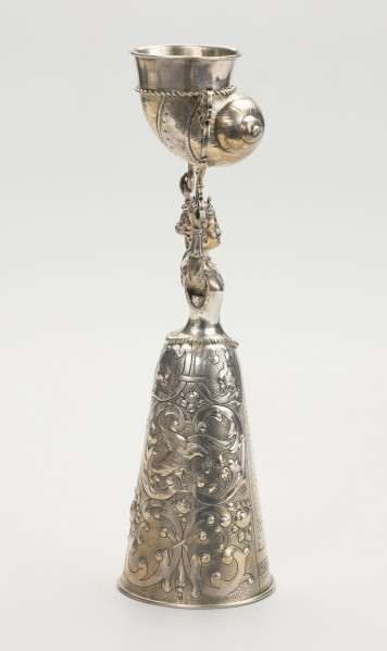 puchar weselny - ujęcie z prawej; Puchar w kształcie młodej kobiety w renesansowej sukni z mocno zasznurowanym staniczkiem i spódnicą o stożkowym kształcie, zdobioną ornamentem schweifwerkowym przechodzącymi w liście akantu, motywami ptaków i kiści owoców. Włosy dziewczyny są spięte z tyłu w koczek, na głowie nosi niewielką koronę z czterema kwiatonami. W rękach wzniesionych nad głową trzyma parę ażurowych wolutowych wsporników, pomiędzy którymi na trzpieniach zamontowany został pucharek w kształcie muszli.