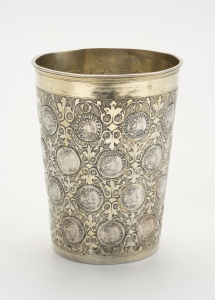 kubek monetowy - ujęcie z przodu; Kubek o konicznym kształcie, rozszerzający się ku górze, z pogrubionym, profilowanym brzegiem. Korpus naczynia wysadzany jest 36 trójgroszami pruskimi Zygmunta I Starego z lat 1534-1540 widocznymi od strony awersu z portretem i tytulatura władcy. Powierzchnię kubka między monetami pokryto symetrycznie zakomponowanym rytowanym ornamentem ze stylizowanej wici roślinnej na matowym, punktowanym tle; przy podstawie oraz przy wylewie ujęty pasami gładkiej powierzchni – szerszym przy wylewie, a węższym przy podstawie.