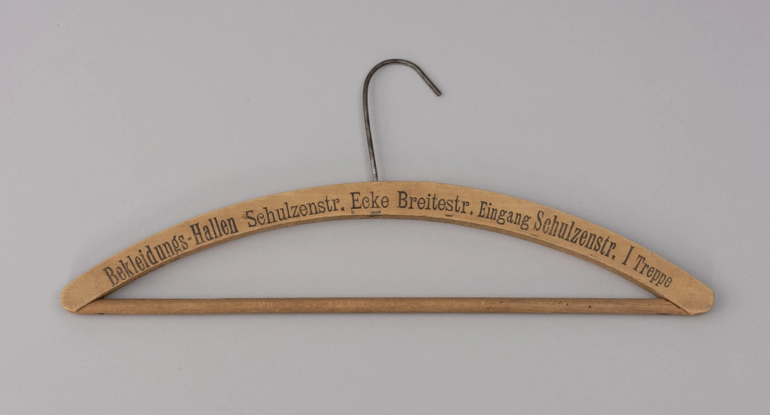 wieszak do szafy - Ujęcie z przodu; Drewniany wieszak ubraniowy z czarnym drukowanym napisem na przodzie BEKLEIDUNGS-HALLEN SCHULZENSTR. ECKE BREITESTR. EINGANG SCHULZENSTR. I TREPPE. Napis w jednej linii, w początkowym fragmencie delikatnie wyblakły. Wieszak zaopatrzony jest w drewnianą poprzeczkę oraz metalowy haczyk. Drewno lakierowane.