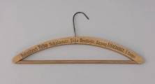 wieszak do szafy - Ujęcie z przodu; Drewniany wieszak ubraniowy z czarnym drukowanym napisem na przodzie BEKLEIDUNGS-HALLEN SCHULZENSTR. ECKE BREITESTR. EINGANG SCHULZENSTR. I TREPPE. Napis w jednej linii, w początkowym fragmencie delikatnie wyblakły. Wieszak zaopatrzony jest w drewnianą poprzeczkę oraz metalowy haczyk. Drewno lakierowane.
