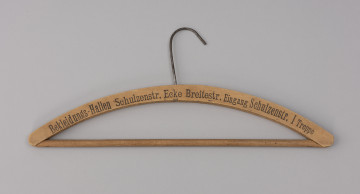 wieszak do szafy - Ujęcie z przodu; Drewniany wieszak ubraniowy z czarnym drukowanym napisem na przodzie BEKLEIDUNGS-HALLEN SCHULZENSTR. ECKE BREITESTR. EINGANG SCHULZENSTR. I TREPPE. Napis w jednej linii, w początkowym fragmencie delikatnie wyblakły. Wieszak zaopatrzony jest w drewnianą poprzeczkę oraz metalowy haczyk. Drewno lakierowane.