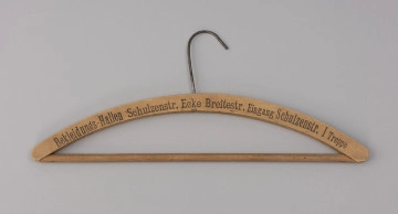 wieszak do szafy - Ujęcie z przodu; Drewniany wieszak ubraniowy z czarnym drukowanym napisem na przodzie BEKLEIDUNGS-HALLEN SCHULZENSTR. ECKE BREITESTR. EINGANG SCHULZENSTR. I TREPPE. Napis w jednej linii, w początkowym fragmencie delikatnie wyblakły. Wieszak zaopatrzony jest w drewnianą poprzeczkę oraz metalowy haczyk. Drewno lakierowane.