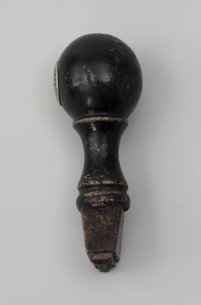 pieczątka firmowa - Ujęcie z boku; Pieczątka z napisem do odbijania, wykonanym minuskułą typu gotyckiego. Napis wycięty w wąskim prostokątnym kawałku tworzywa sztucznego (prawdopodobnie linoleum) i przyklejony do drewnianego uchwytu zakończonego okrągłą główką, na której z lewej strony przymocowano za pomocą nitu metalowy znak firmowy okrągły. Od główki odchodzi wąska szyjka, do której przytwierdzony jest element o przekroju w kształcie trapezu. Drewniana część tłocząca zabarwiona czarnym lakierem.