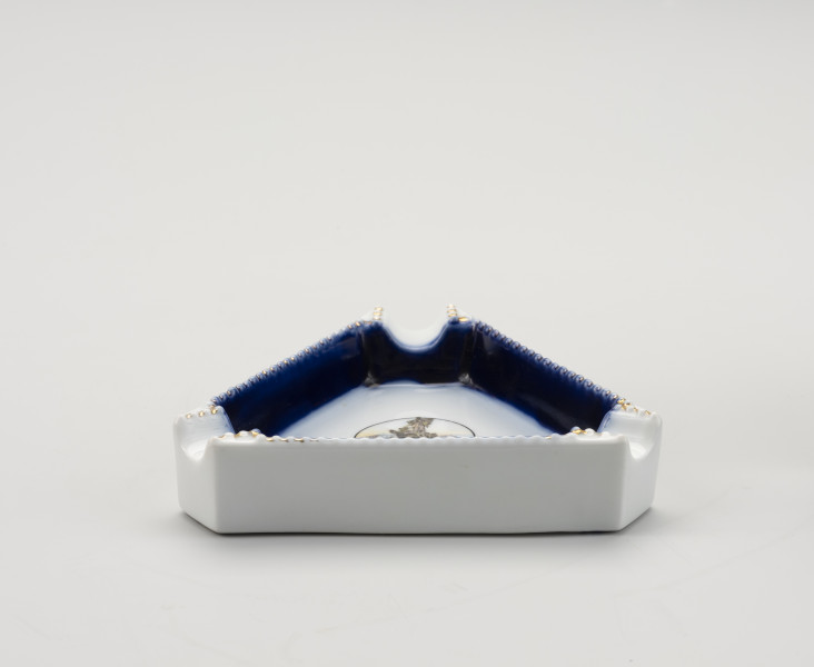 popielniczka pamiątkowa z wyobrażeniem Sediny - Ujęcie z przodu; Porcelanowa popielniczka w kształcie równobocznego trójkąta. Biała, z granatowym obrzeżem na widocznych dwóch ściankach wewnątrz. W każdym rogu wyżłobione miejsce na papierosa/cygaro. Brzeżki górne granulkowe, biało-złote. Wewnątrz popielnicy, na trójkątnym dnie, w środkowej części kółko, a w nim kolorowy rysunek.