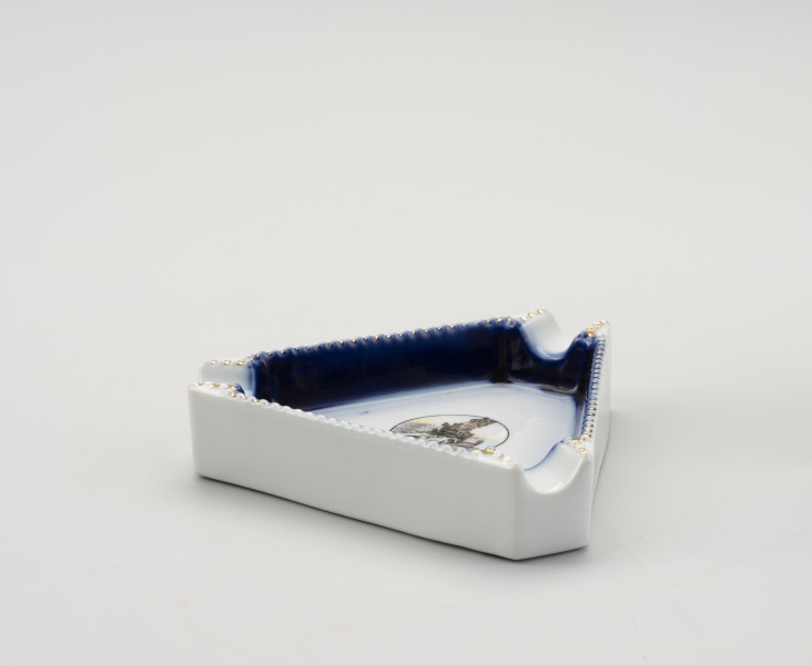 popielniczka pamiątkowa z wyobrażeniem Sediny - Ujęcie z boku; Porcelanowa popielniczka w kształcie równobocznego trójkąta. Biała, z granatowym obrzeżem na dwóch widocznych ściankach wewnątrz. W każdym rogu wyżłobione miejsce na papierosa/cygaro. Brzeżki górne granulkowe, biało-złote. Wewnątrz popielnicy, na trójkątnym dnie, w środkowej części kółko, a w nim kolorowy rysunek.