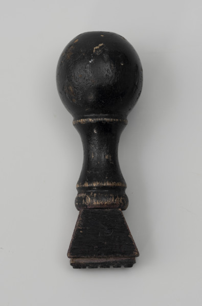 pieczątka firmowa - Ujęcie z tyłu; Pieczątka z napisem do odbijania, wykonanym minuskułą typu gotyckiego. Napis wycięty w wąskim prostokątnym kawałku tworzywa sztucznego (prawdopodobnie linoleum) i przyklejony do drewnianego uchwytu zakończonego okrągłą główką. Od główki odchodzi wąska szyjka, do której przytwierdzony jest element o przekroju w kształcie trapezu. Drewniana część tłocząca zabarwiona czarnym lakierem.