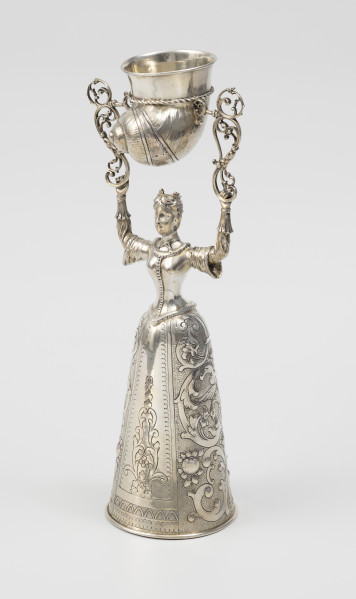 puchar weselny - ujęcie ze skosu z prawej; Puchar w kształcie młodej kobiety w renesansowej sukni z mocno zasznurowanym staniczkiem i spódnicą o stożkowym kształcie, zdobioną ornamentem schweifwerkowym przechodzącymi w liście akantu, motywami ptaków i kiści owoców. Na głowie nosi niewielką koronę z czterema kwiatonami. W rękach wzniesionych nad głową trzyma parę ażurowych wolutowych wsporników, pomiędzy którymi na trzpieniach zamontowany został pucharek w kształcie muszli.