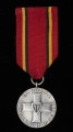 Medal za Udział w Walkach o Berlin - ujęcie z przodu; Oznaką medalu jest wykonany z metalu srebrzonego i oksydowanego krążek, na którego awersie umieszczony jest centralnie wizerunek orderu Krzyża Grunwaldu otoczony na krawędzi koła napisem: ZA UDZIAŁ W WALKACH O BERLIN. Medal zawieszony jest na czerwonej wstążce, z dwoma czarno-żółtymi paskami po bokach. Zawieszenie z przylutowanego uszka z kółeczkiem.