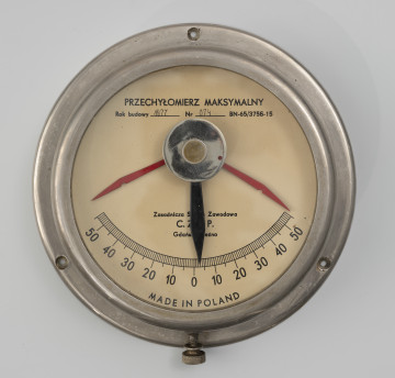 przechyłomierz maksymalny - Ujęcie z przodu; Przechyłomierz o metalowym korpusie ze szklanym zabezpieczeniem tarczy. Średnica górnej części wynosi 18 cm. Rant zaokrąglony. Podstawę stanowi kołnierz o średnicy 21 cm, na którym znajdują się 3 otwory na śruby. W dolnej części białej tarczy skala kątowa 100-stopniowa w zakresach 50° – 0° – 50°. Powyżej środka oś z czarną wskazówką zawieszoną wewnątrz na urządzeniu balastowym. Na osi wskazówki dwa czerwone ograniczniki kąta wychylenia i pokrętło do ustawiania ograniczników. Na tarczy, w centrum, poniżej wskazówki sygnatura producenta Zasadnicza Szkoła Zawodowa C. Z. S. P. Gdańsk - Brzeźno. Powyżej wskazówki napis PRZECHYŁOMIERZ MAKSYMALNY, rok budowy 1977, nr 074, BN - 65, 3756 - 15. W dolnej części przy krawędzi tarczy napis Made in Poland.