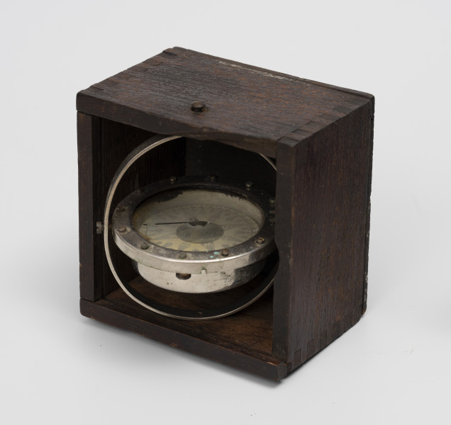 Rybacki kompas morski - ujęcie z góru; Kociołek kompasu mokrego zainstalowany na zawieszeniu Cardana w drewnianym pudełku. Róża kompasowa z podziałką rumbową i stopniową (dziesiąta cześć stopni w numeracji od 0 do 35 stopni). Na szybie kociołka namalowana czarna kreska kursowa. Zawieszenie Cardana wyposażone w owalny pierścień o przekątnych długości 8 i 8,5 cm. Kociołek, pierścień azymutalny i pierścień zawieszenia Cardana niklowane. Brak zasuwanej pokrywki pudełka.