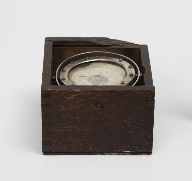 Rybacki kompas morski - ujęcie z boku; Kociołek kompasu mokrego zainstalowany na zawieszeniu Cardana w drewnianym pudełku. Na szybie kociołka namalowana czarna kreska kursowa. Zawieszenie Cardana wyposażone w owalny pierścień o przekątnych długości 8 i 8,5 cm. Kociołek, pierścień azymutalny i pierścień zawieszenia Cardana niklowane. Brak zasuwanej pokrywki pudełka.
