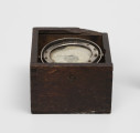 Rybacki kompas morski - ujęcie z boku; Kociołek kompasu mokrego zainstalowany na zawieszeniu Cardana w drewnianym pudełku. Na szybie kociołka namalowana czarna kreska kursowa. Zawieszenie Cardana wyposażone w owalny pierścień o przekątnych długości 8 i 8,5 cm. Kociołek, pierścień azymutalny i pierścień zawieszenia Cardana niklowane. Brak zasuwanej pokrywki pudełka.