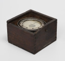 Rybacki kompas morski - ujęcie z boku; Kociołek kompasu mokrego zainstalowany na zawieszeniu Cardana w drewnianym pudełku. Na szybie kociołka namalowana czarna kreska kursowa. Zawieszenie Cardana wyposażone w owalny pierścień o przekątnych długości 8 i 8,5 cm. Kociołek, pierścień azymutalny i pierścień zawieszenia Cardana niklowane. Brak zasuwanej pokrywki pudełka.