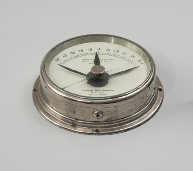 przechyłomierz okrętowy LO 55 - Ujęcie z dołu; Okrągły przechyłomierz okrętowy LO 55 w metalowej, niklowanej puszce o średnicy 19 cm i kołnierzem z otworami do przykręcenia na ścianę, wysokości 5 cm. Na białej tarczy u góry 100-stopiowa skala w zakresach 50° - 0° - 50°. Na tarczy w centrum i u dołu opis PRZECHYŁOMIERZ LO 55, rok budowy 196.., nr 072, Zasadnicza Szkoła Zawodowa C. Z. S. P., Gdańsk - Brzeźno, Made in Poland. Poniżej środka tarczy mocowanie wskaźnika przechyłu z przeciwwagą, utrzymującą wskaźnik w pozycji pionowej, wewnątrz pod tarczą. Po bokach wskaźnika dwa ograniczniki wskazań przechyłu. Na szybie przykrywającej tarczę pokrętło do regulacji wskazówek maksymalnego przechyłu.