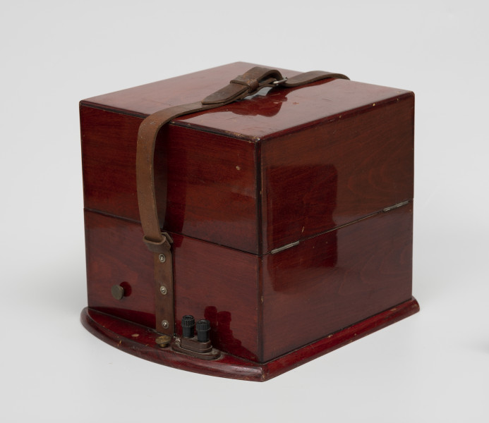 chronometr morski Gost - Ujęcie ze skosu; Skrzynka drewniana kwadratowa w kolorze ciemnej wiśni, lakierowana na wysoki połysk. Do boków skrzynki przytwierdzone zostały pasy skórzane do transportu, a do podstawy, po lewej stronie przykręcone uchwyty do mocowania kabli.