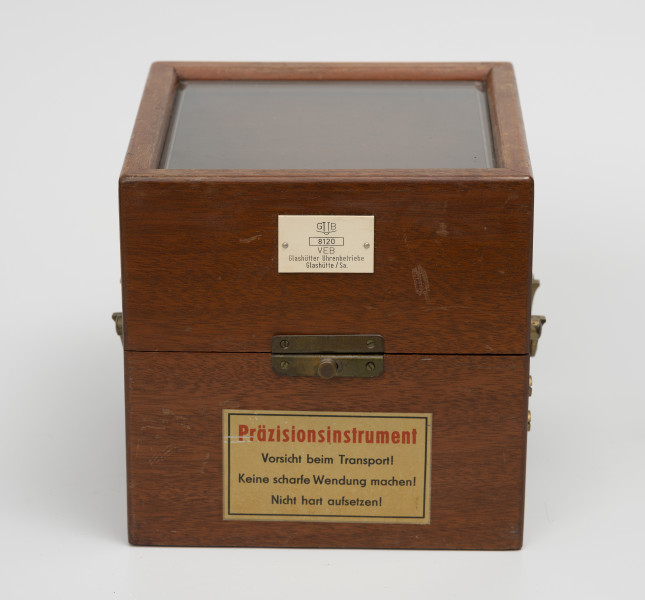 Chronometr na zawieszeniu Cardana - ujęcie z przodu; Chronometr w drewnianej skrzynce z kluczem. Skrzynka drewniana z mahoniu, lakierowana, matowa. Wieczko skrzynki z szybą fazowaną. Powyżej okucia biała, plastikowa tabliczka znamionowa o wymiarach 4,3x2,7 cm, poniżej nalepka informacyjna.