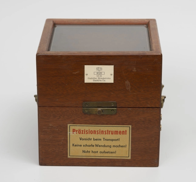 Chronometr na zawieszeniu Cardana - ujęcie z przodu; Chronometr w drewnianej skrzynce z kluczem. Skrzynka drewniana z mahoniu, lakierowana, matowa. Wieczko skrzynki z szybą fazowaną. Powyżej okucia biała, plastikowa tabliczka znamionowa o wymiarach 4,3x2,7 cm, poniżej nalepka informacyjna.