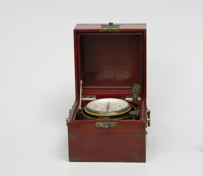 chronometr morski Gost - Ujęcie z przodu; Otwarta skrzynka drewniana kwadratowa w kolorze ciemnej wiśni, lakierowana na wysoki połysk. Na bokach uchwyty z mosiądzu do przenoszenia. Łączenia boczne skrzynki wzmocnione 12 mosiężnymi kątownikami. Skrzynka jest trójdzielna, zamykana na zamki zatrzaskowe. Wewnątrz na zawieszeniu Cardana osadzony jest chronometr, blokowany metalową blokadą na mosiężnej śrubie. Biały metalowy cyferblat z arabskimi, przetłoczonymi cyframi w kolorze czarnym. Część kociołka zamykana mosiężnym pierścieniem z szybą. W górnej części tarczy mała okrągła skala do odmierzania 56-godzinnego czasu pracy urządzenia z wartościami co 8 godzin.