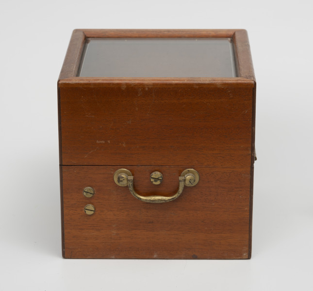 Chronometr na zawieszeniu Cardana - ujęcie z boku z prawej; Chronometr w drewnianej skrzynce z kluczem. Skrzynka drewniana z mahoniu, lakierowana, matowa. Wieczko skrzynki z szybą fazowaną. Na bocznej ścianie widoczne mosiężne śruby.