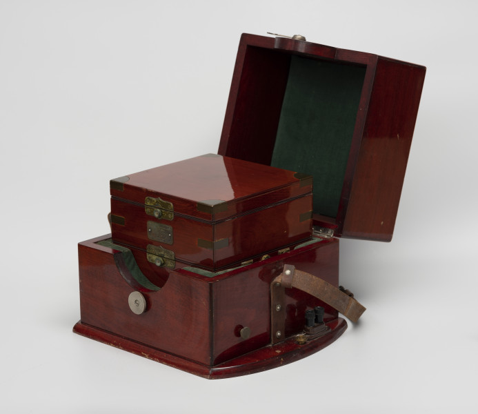 chronometr morski Gost - Ujęcie ze skosu z prawej; Otwarta skrzynka drewniana kwadratowa w kolorze ciemnej wiśni, lakierowana na wysoki połysk o wymiarach 25x22x25 cm, z wnętrzem wyściełanym zielonym materiałem. Do boków skrzynki przytwierdzone zostały pasy skórzane do transportu, a do podstawy, po prawej stronie przykręcone uchwyty do mocowania kabli. Wewnątrz kolejna skrzynka drewniana kwadratowa w kolorze ciemnej wiśni, lakierowana na wysoki połysk. Łączenia boczne wewnętrznej skrzynki wzmocnione 12 mosiężnymi kątownikami. Od czoła tabliczka mosiężna o wymiarach 4,8x2,5 cm o treści Moskowskoj Cziasowoj Zawod im S.M.Kirowa Morskoj Chronometr Gost 8916-58. Skrzynka jest trójdzielna, zamykana na zamki zatrzaskowe.