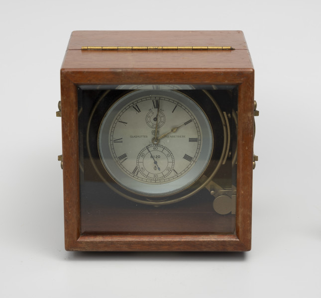 Chronometr na zawieszeniu Cardana - ujęcie z góry; Chronometr w drewnianej skrzynce. Skrzynka drewniana z mahoniu, lakierowana, matowa. Wieczko skrzynki z szybą fazowaną. Górna i dolna część skrzynki połączona zawiasem pasowym. Cyferblat chronometru biały z czarnymi oznaczeniami. Godziny w rzymskim zapisie liczbowym, obwiedzione podziałka minutową. Cyfry tarczy sekundowej i czasu pracy arabskie. Wskazówki, godzinowa w formie strzałki i prosta minutowa, wykonane z mosiądzu. Wskazówka sekundnikowa i czasu pracy z pomalowanego na czarno metalu. Podziałka wskaźnika godzin czasu pracy w formie niepełnego okręgu, umieszczony pod rzymską godziną XII i wyskalowany od 0 do 56, przy czym wartość 0 z oznaczeniem AUF, a wartość 56 z oznaczeniem AB.