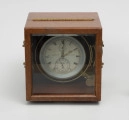 Chronometr na zawieszeniu Cardana - ujęcie z góry; Chronometr w drewnianej skrzynce. Skrzynka drewniana z mahoniu, lakierowana, matowa. Wieczko skrzynki z szybą fazowaną. Górna i dolna część skrzynki połączona zawiasem pasowym. Cyferblat chronometru biały z czarnymi oznaczeniami. Godziny w rzymskim zapisie liczbowym, obwiedzione podziałka minutową. Cyfry tarczy sekundowej i czasu pracy arabskie. Wskazówki, godzinowa w formie strzałki i prosta minutowa, wykonane z mosiądzu. Wskazówka sekundnikowa i czasu pracy z pomalowanego na czarno metalu. Podziałka wskaźnika godzin czasu pracy w formie niepełnego okręgu, umieszczony pod rzymską godziną XII i wyskalowany od 0 do 56, przy czym wartość 0 z oznaczeniem AUF, a wartość 56 z oznaczeniem AB.