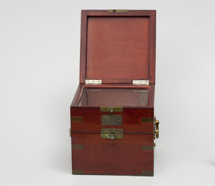 chronometr morski Gost - Ujęcie z przodu; Otwarta skrzynka drewniana kwadratowa w kolorze ciemnej wiśni, lakierowana na wysoki połysk. Na bokach uchwyty z mosiądzu do przenoszenia. Łączenia boczne skrzynki wzmocnione 12 mosiężnymi kątownikami. Od czoła tabliczka mosiężna o wymiarach 4,8x2,5 cm o treści Moskowskoj Cziasowoj Zawod im S.M.Kirowa Morskoj Chronometr Gost 8916-58. Skrzynka jest trójdzielna, zamykana na zamki zatrzaskowe; środkowa pokrywa posiada szkło umożliwiające obserwację chronometru bez otwierania wieczka.