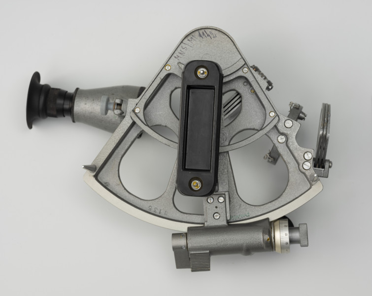 sekstant SNO-T w skrzynce z akcesoriami i atestami - Ujęcie z tyłu; Sekstant z lunetką i przeziernikiem. Odlewana, metalowa rama z jednym bokiem trójkąta w kształcie wycinka koła łączona wewnętrznie kratownicą w kolorze metalizowanym o wymiarach18,5x18,5x23 cm. Od przodu umocowane ruchome ramię, alidada o długości23 cm. Ramię alidady porusza się po wyskalowanym boku trójkąta, limbusie. Na końcu ramienia śruba ślimakowa w obudowie. Przy zakończeniu obudowy znajduje się śruba zakończona bębenkiem z podziałką minutową. Śruba ślimakowa zazębia się z zębatką naciętą na krawędzi ramy. Na końcu obudowy znajduje się uchwyt wyłączający śrubę. Na alidadzie wybito literę E z numerem 135. Za lusterkiem małym z prawej trzy przesłony o różnym stopniu przyciemniania. Z przodu widoczny czarny, plastikowy uchwyt na dłoń i dwie nóżki służące za podstawę. Lunetka z lewej o długości 11,2 cm. przeziernik o długości 17,5 cm.