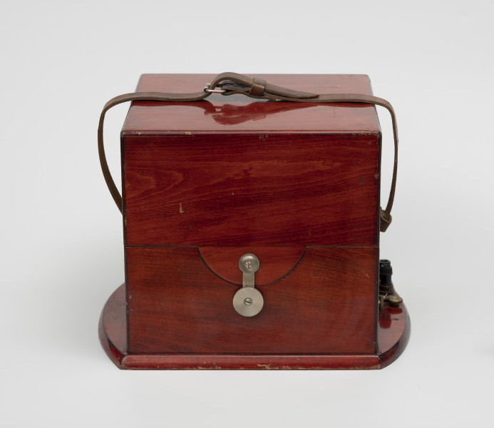 chronometr morski Gost - Ujęcie z przodu; Skrzynka drewniana kwadratowa w kolorze ciemnej wiśni, lakierowana na wysoki połysk. Do boków skrzynki przytwierdzone zostały pasy skórzane do transportu, a do podstawy, po prawej stronie przykręcone uchwyty do mocowania kabli.