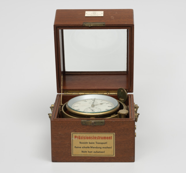 Chronometr na zawieszeniu Cardana - ujęcie z przodu; Chronometr w drewnianej skrzynce z kluczem. Skrzynka drewniana z mahoniu, lakierowana. Wieczko skrzynki z szybą fazowaną; boki podstawy skrzynki zaopatrzone w mosiężne ucha. Od frontu zamknięcie typu zapinka w formie okucia, na wieku płaskie, u podstawy zaopatrzone w trzpień. Powyżej okucia biała, plastikowa tabliczka znamionowa o wymiarach 4,3x2,7 cm, poniżej nalepka informacyjna. Z lewej strony dolnej części skrzynki mosiężna, dokręcana śrubami prowadnica, zabezpieczającą wieko przed zamknięciem i wyłamaniem. Krawędzie zamknięcia skrzynki z frezowaniem typu pióro-wpust, służące uszczelnieniu. 
Cyferblat chronometru biały z czarnymi oznaczeniami. Godziny w rzymskim zapisie liczbowym, obwiedzione podziałka minutową. Cyfry tarczy sekundowej i czasu pracy arabskie. Wskazówki, godzinowa w formie strzałki i prosta minutowa, wykonane z mosiądzu. Wskazówka sekundnikowa i czasu pracy z pomalowanego na czarno metalu. Podziałka wskaźnika godzin czasu pracy w formie niepełnego okręgu, umieszczony pod rzymską godziną XII. Mosiężny klucz o okrągłym uchwycie na dłoń w górnej części posiada otwór.
